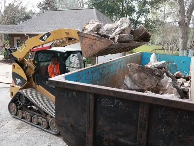 Cat track loader tipping a bucket of broken concrete into a roll-off dumpster in a residential driveway during a house demolition.