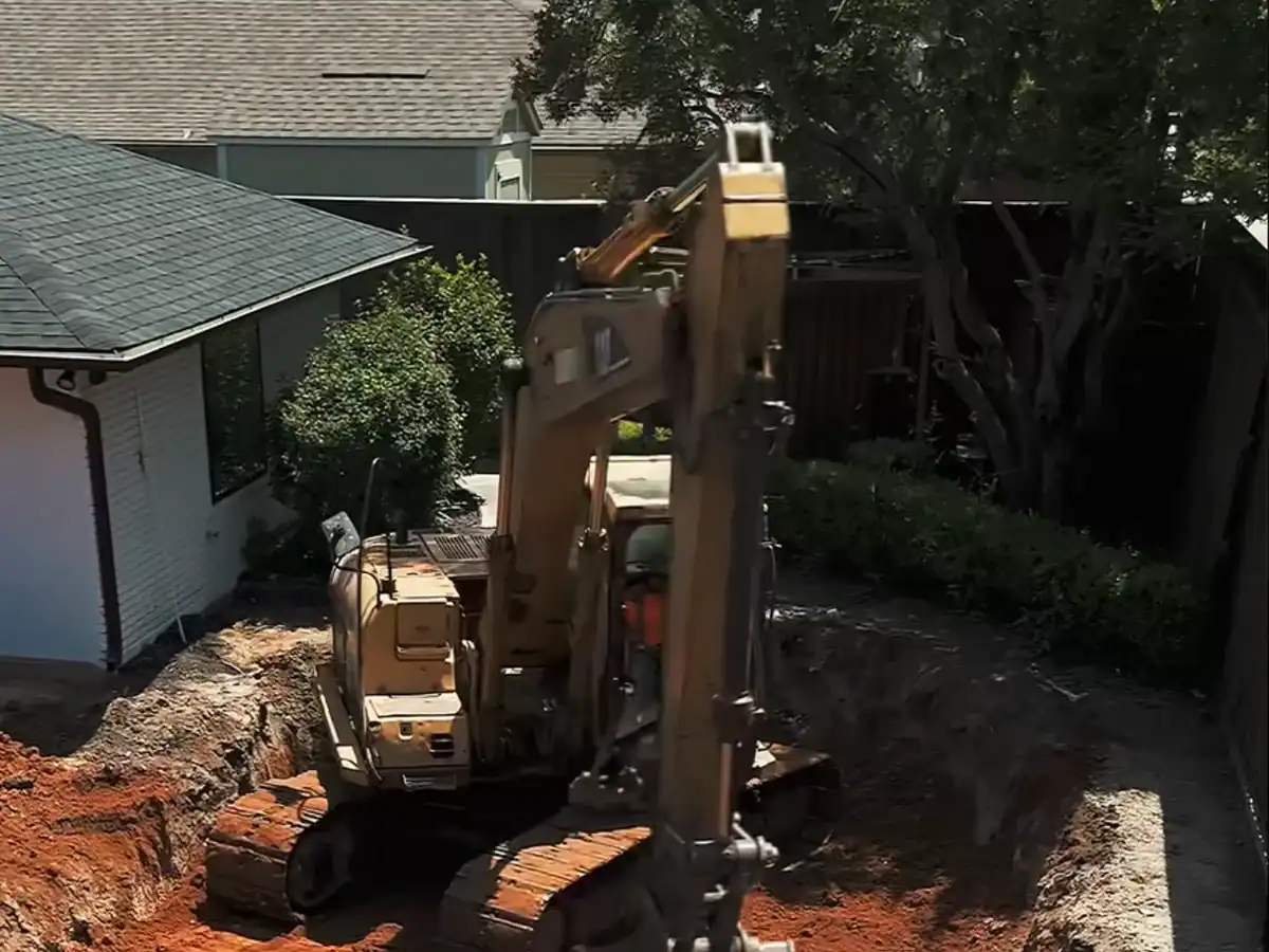 Compact excavator working in the narrow side yard between a house and a wood fence, with the lot cleared down to bare soil.